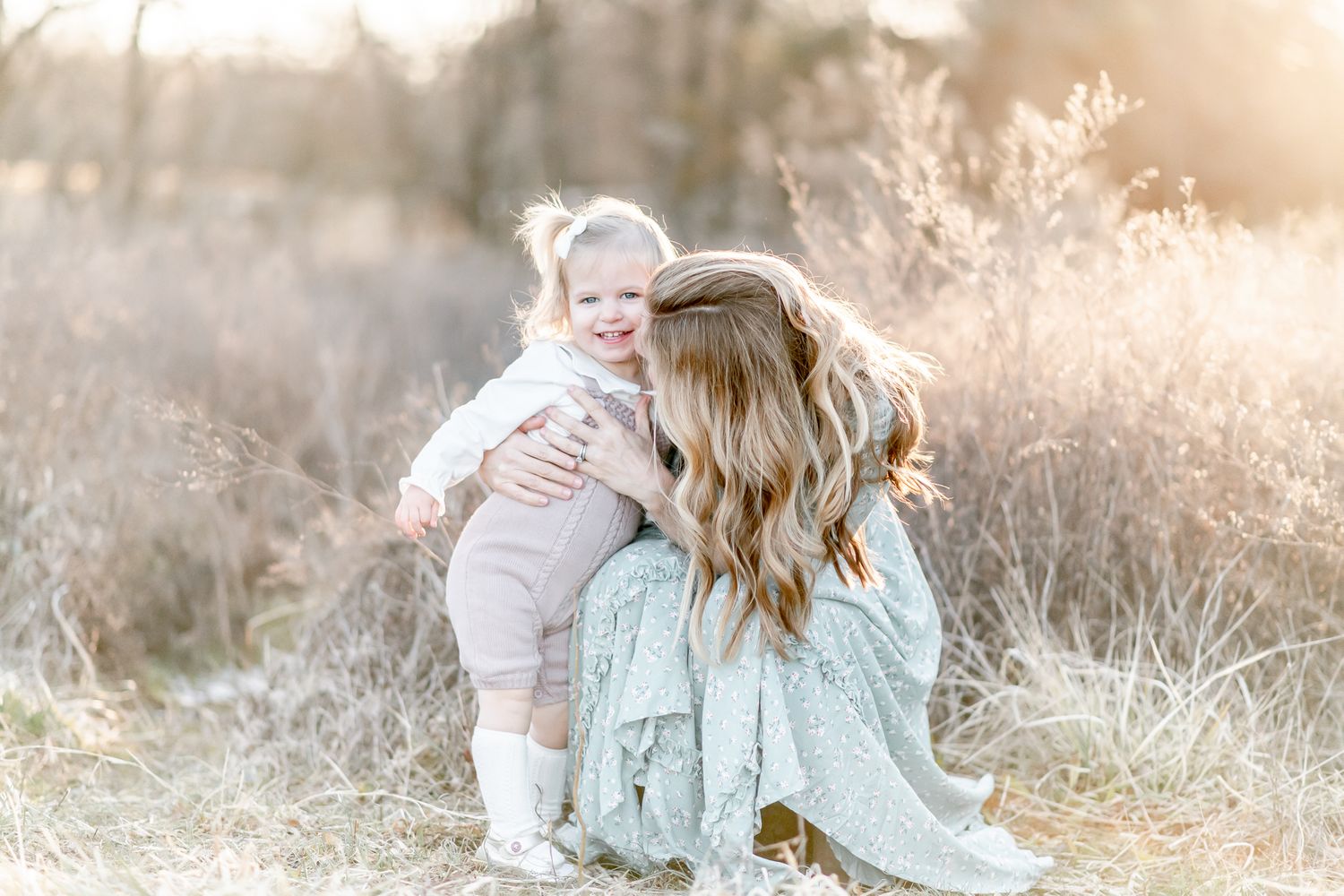 Mom snuggles with toddler girl in a field at sunset