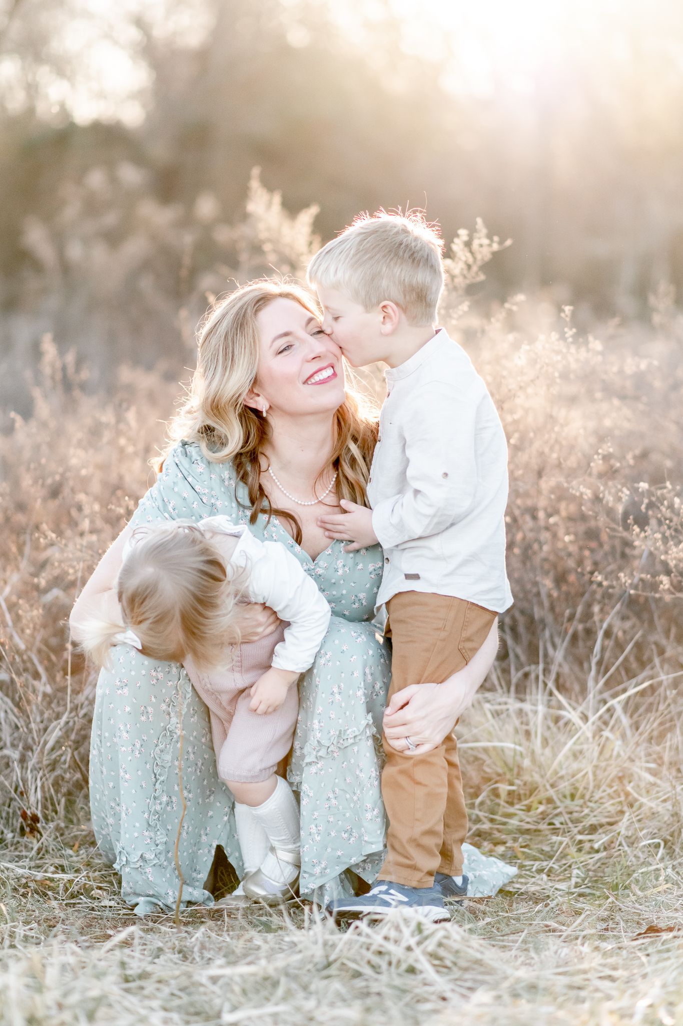 Son kisses mother as she kneels down and also holds toddler