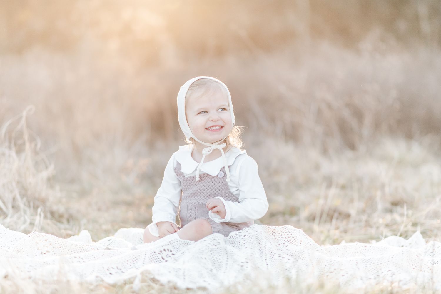 Baby girl in a white Briar Baby Bonnet sits on a blanket in a field at sunset