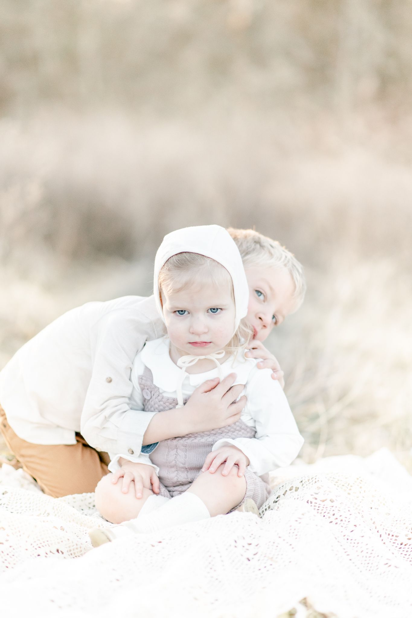 Big brother sneaks a hug behind his baby sister sitting on a blanket