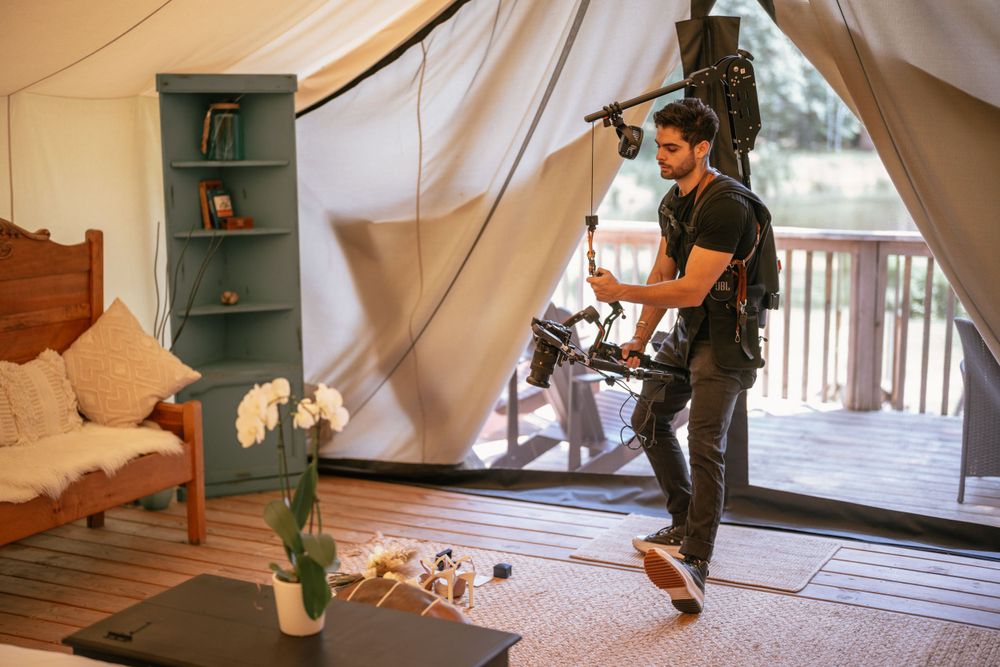 Videographer with professional camera equipment films inside a cozy glamping tent with boho decor and natural lighting.