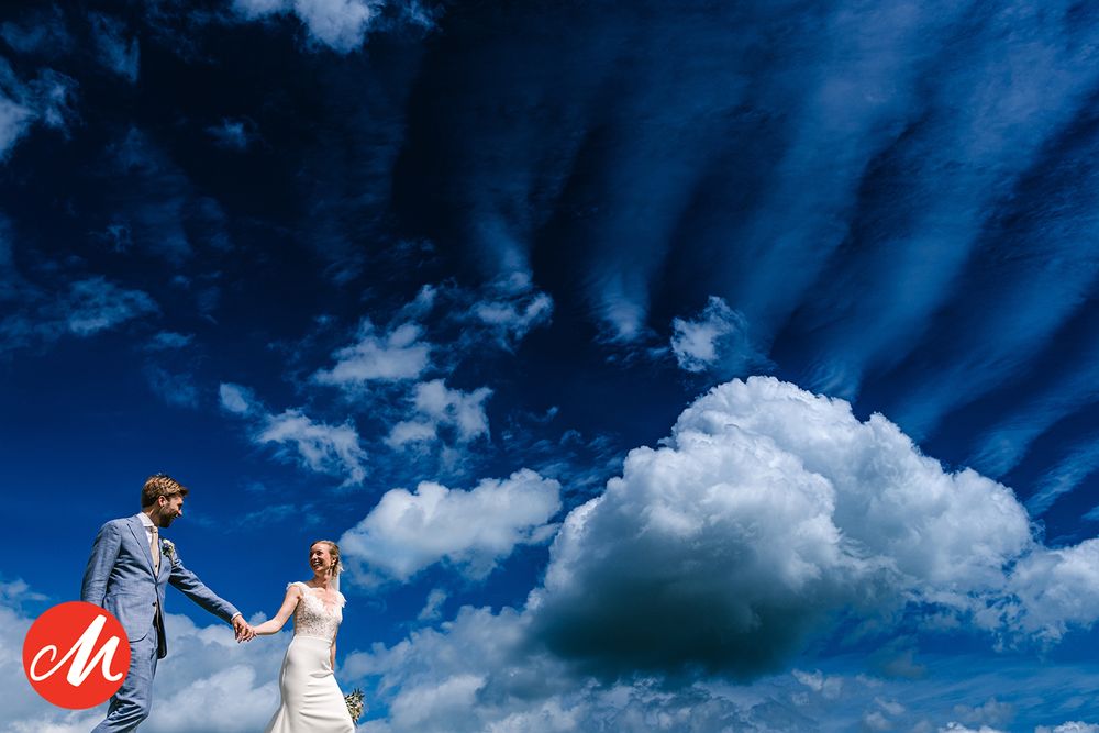 A couple in wedding attire hold hands against a dramatic blue sky with sunbeams and white puffy clouds.