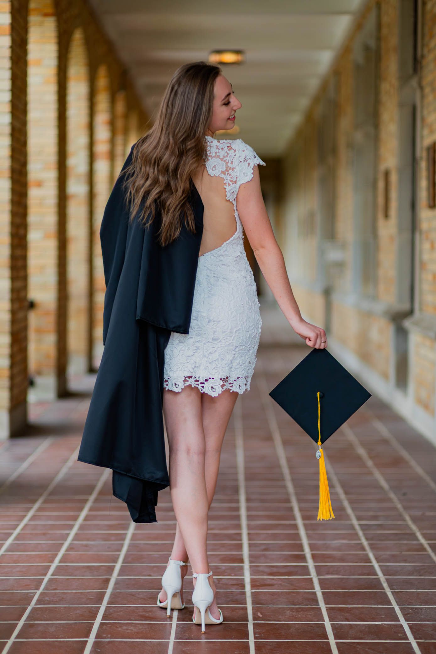 Graduate in white lace dress holding cap while walking through brick hallway.