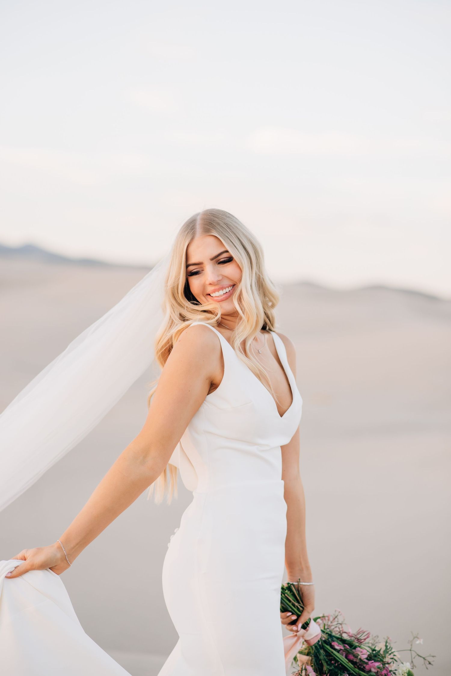 A bride in a fitted white wedding dress poses with her veil flowing in the wind against a desert landscape at sunset.