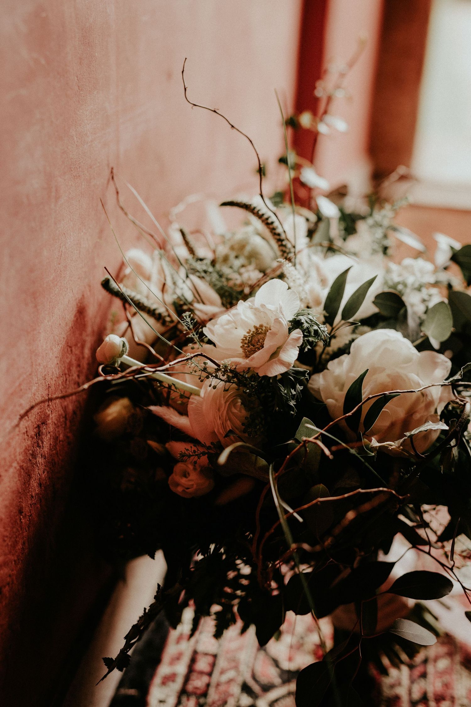 A moody floral arrangement with white roses and wild branches captured in dramatic warm lighting against a textured wall.