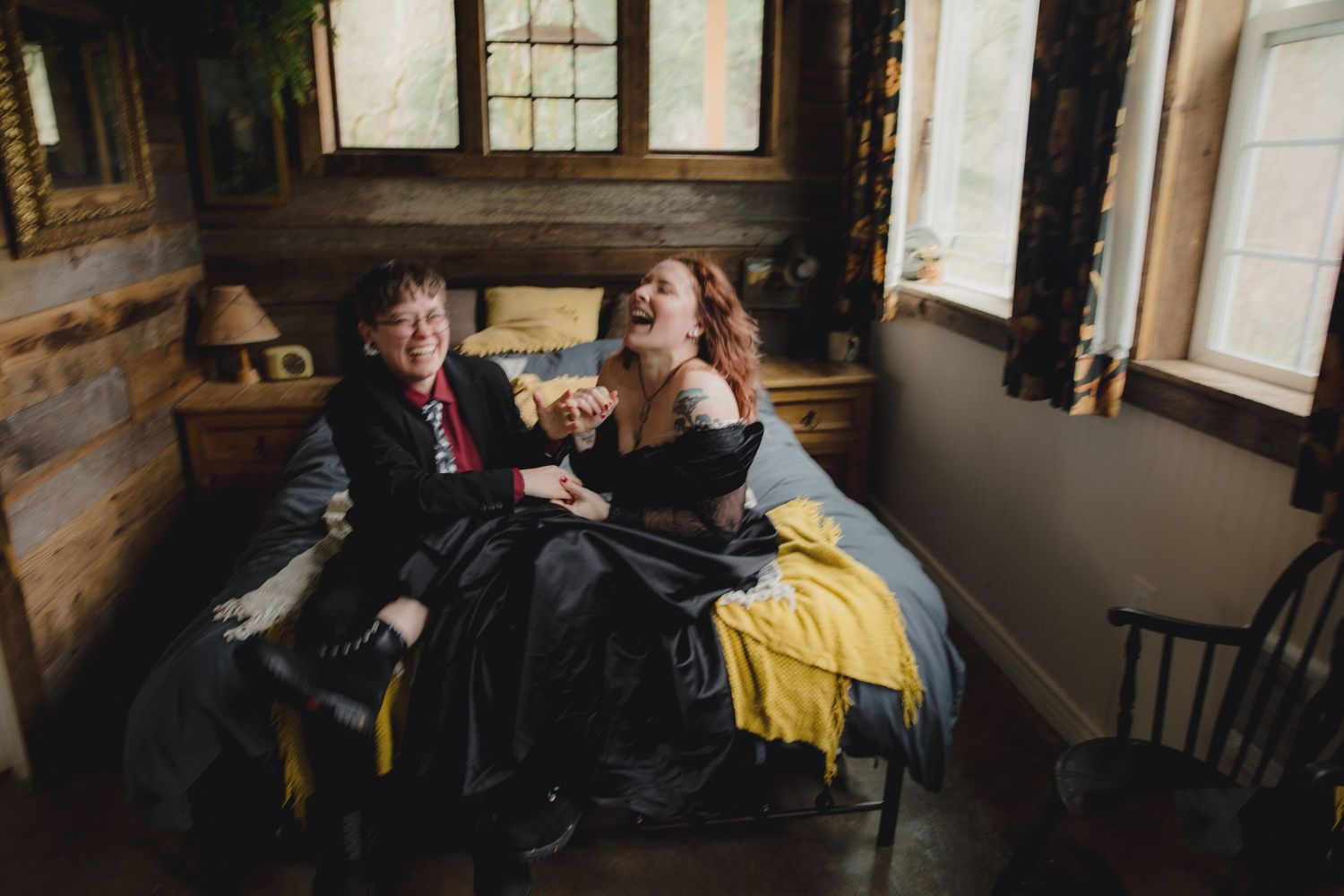 Two people in period clothing share a laugh while sitting on a bed in a rustic cabin with wooden walls and windows.