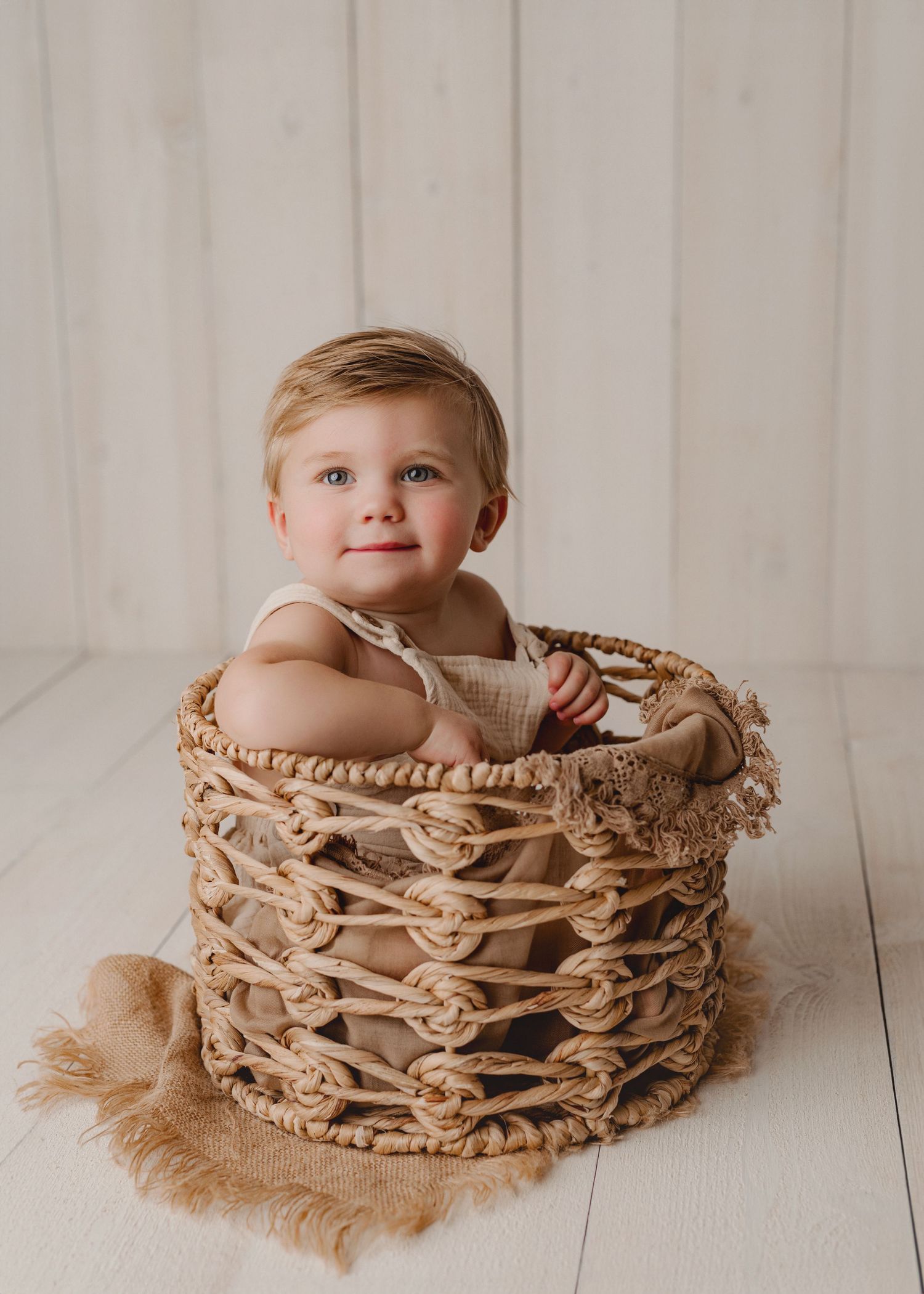 A small child sits in a woven beige macrame basket on a light wooden floor during a photography session.