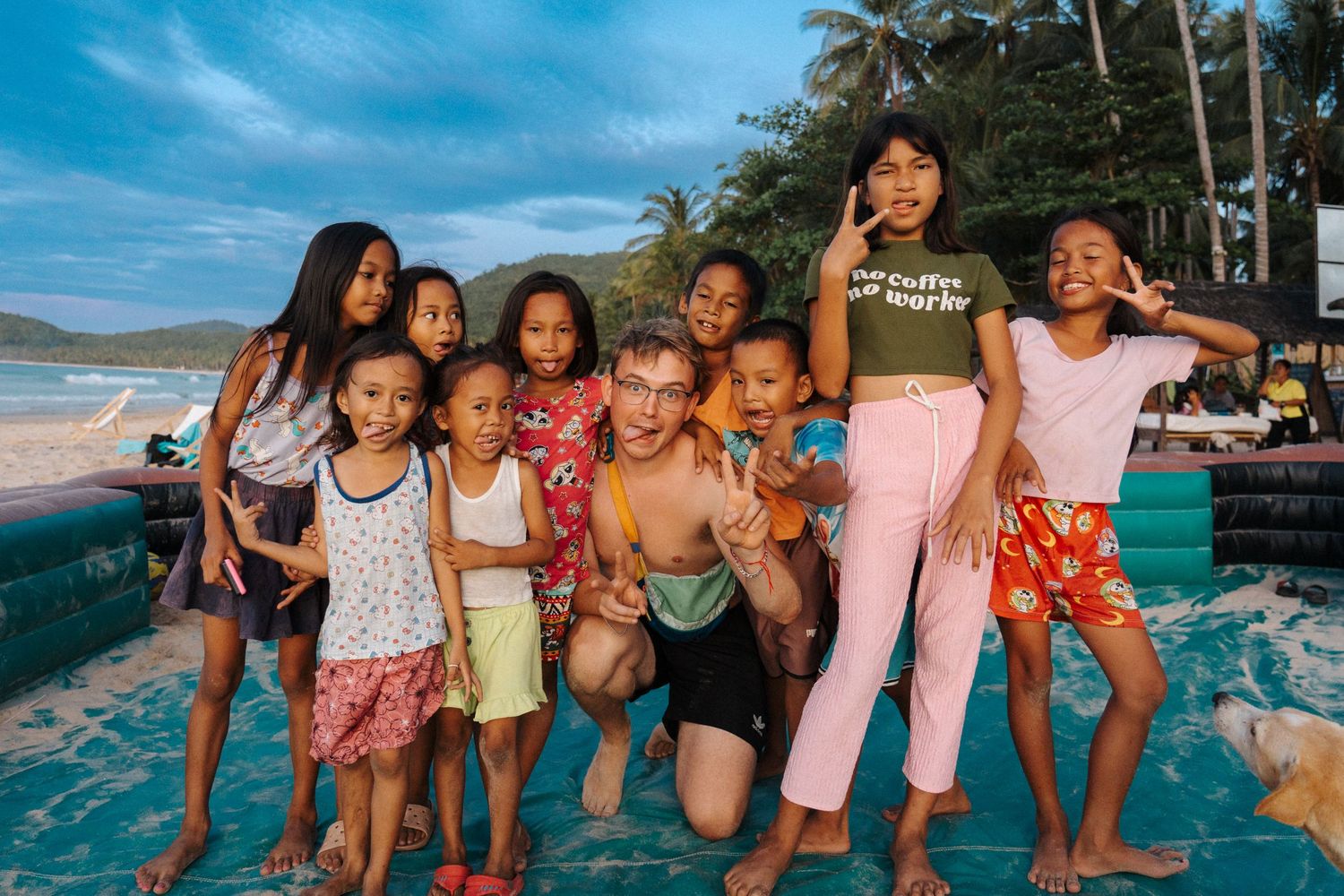 A group of children smile and pose together on a tropical beach with palm trees and blue sky in the background.