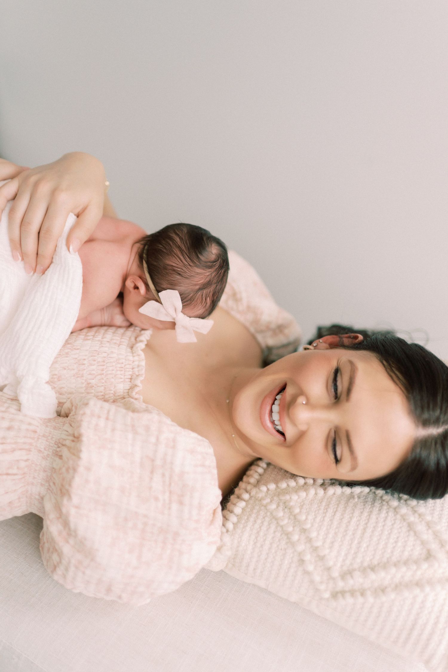 Mother and baby lie peacefully together on white bedding wearing soft pink sweaters.