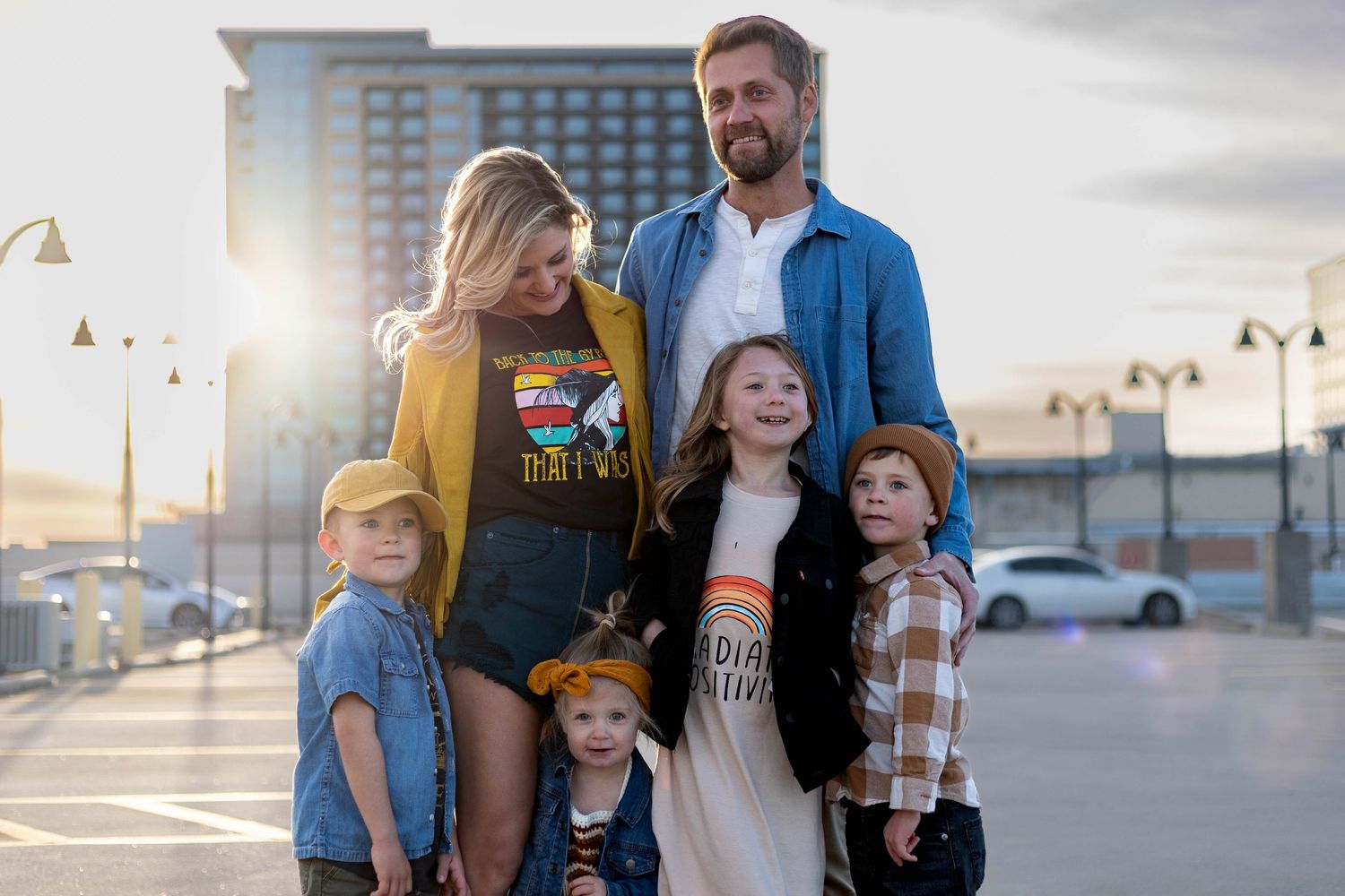 A family of five stands together outdoors at sunset with a city backdrop and lamp posts illuminated behind them.