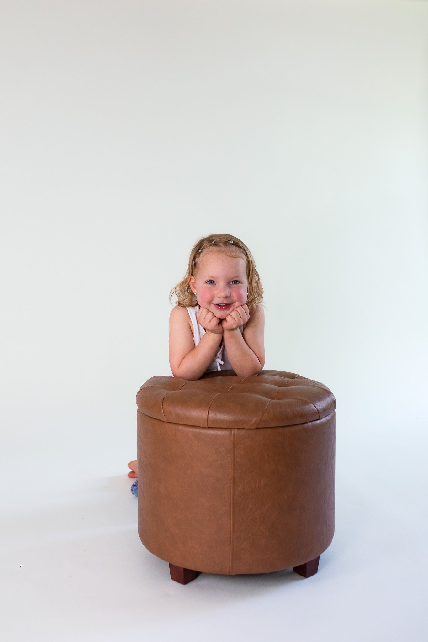 A child rests their arms and chin on a round brown leather ottoman against a white background.