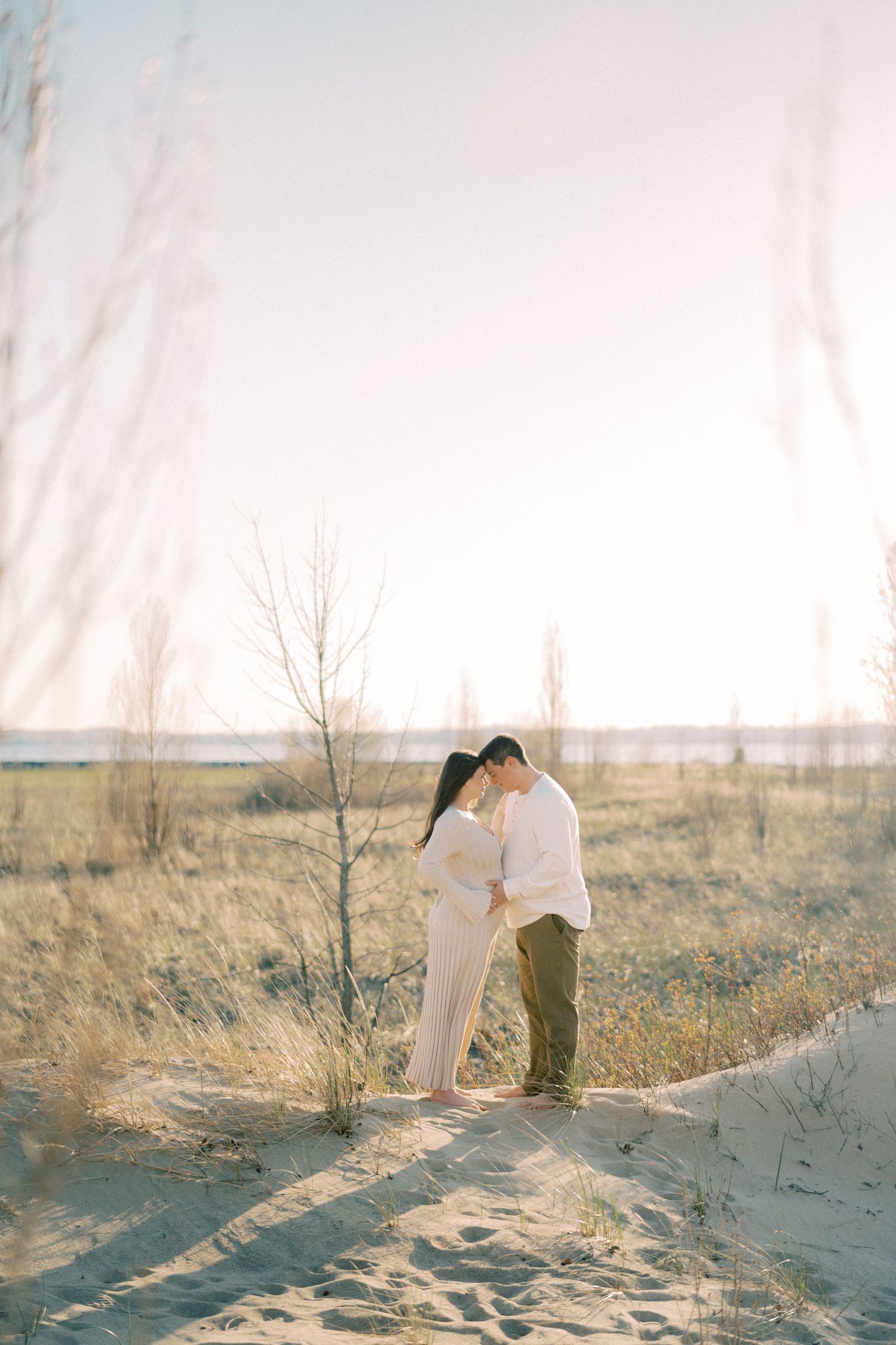 A couple embraces on sandy dunes with bare trees and a misty landscape in the background during golden hour.
