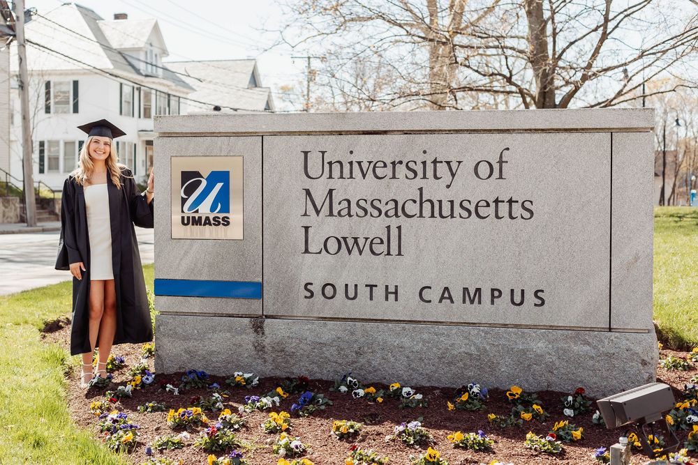 A graduation photo at the University of Massachusetts Lowell South Campus entrance sign with spring flowers blooming.
