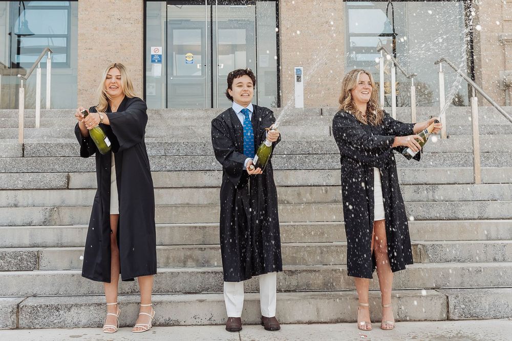 Three graduates celebrating with champagne bottles on stone steps while wearing black graduation gowns.