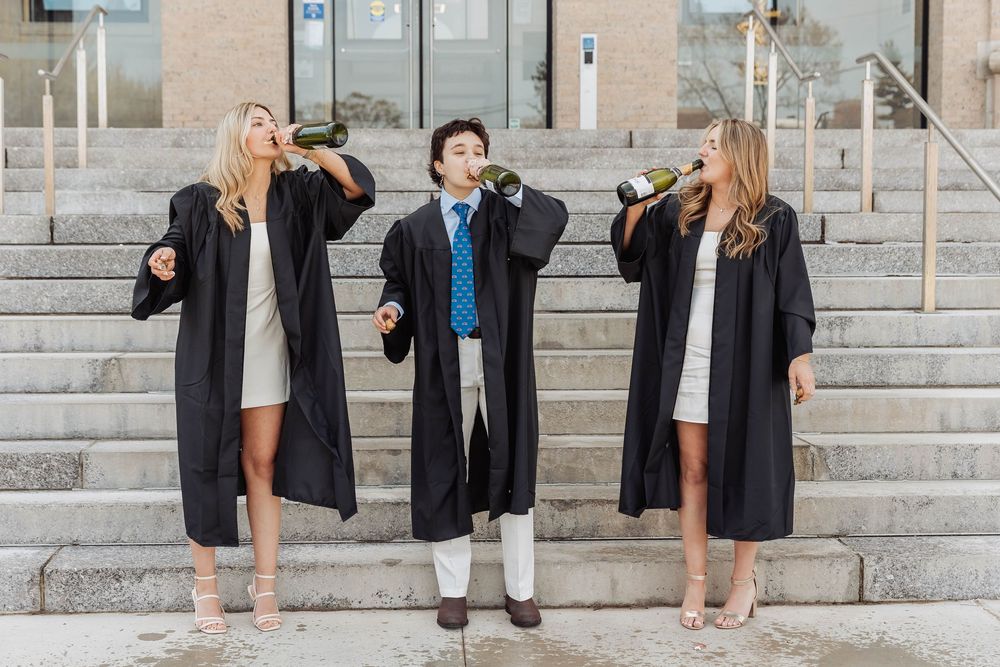 Three graduates in black graduation gowns stand on stone steps celebrating together with enthusiastic gestures.