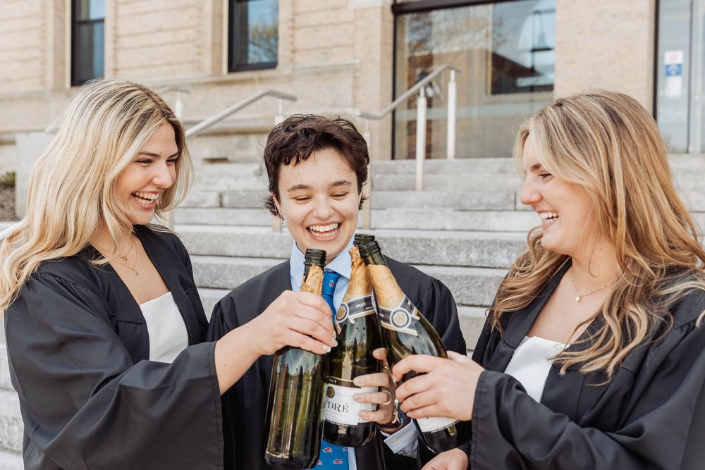 Three graduates in caps and gowns celebrate with a champagne toast on stone steps of a building.