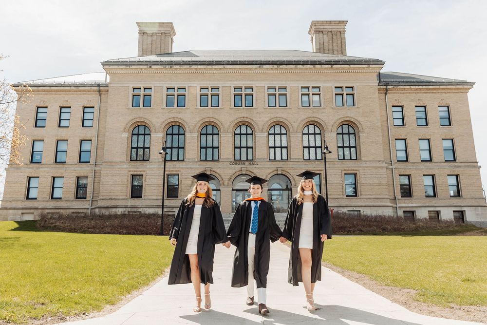 Graduates walk together in front of a grand stone academic building with tall windows on a sunny day.