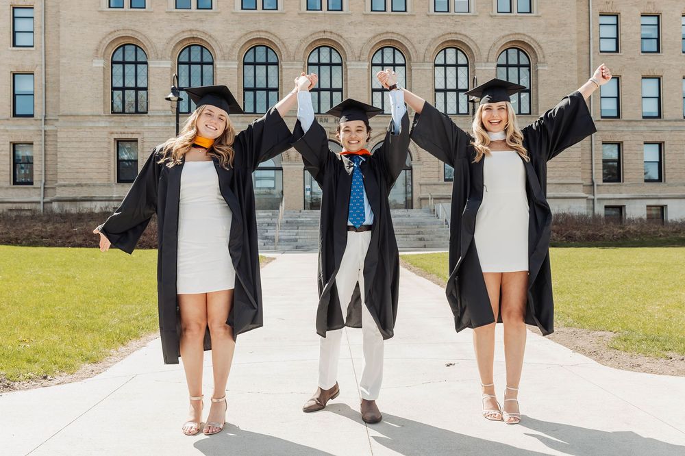 Three graduates in caps and gowns celebrate joyfully with raised arms on a college campus lawn.
