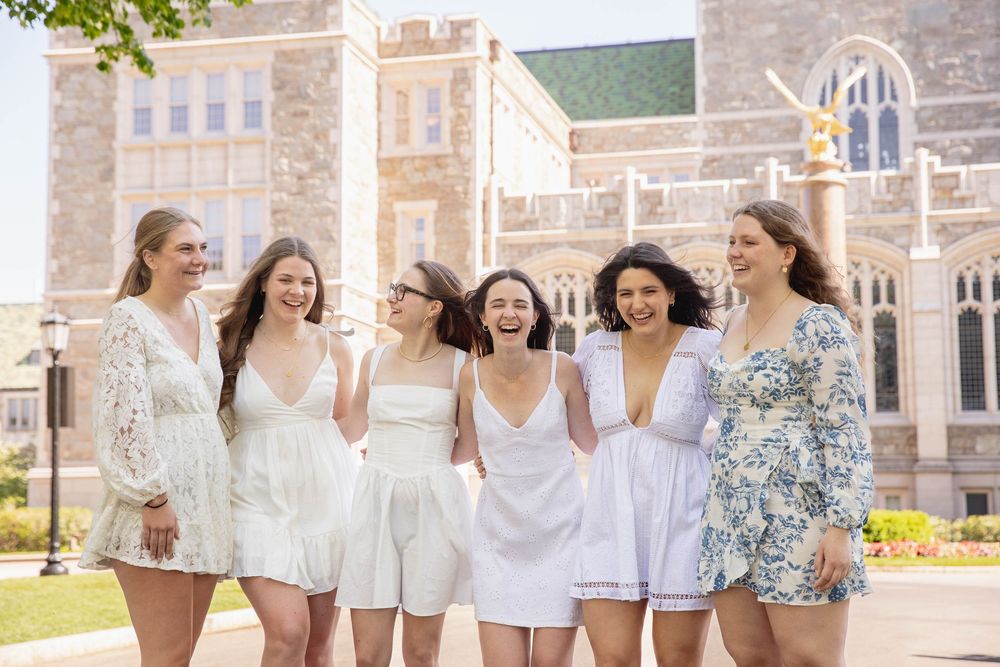 A group of friends wearing light summer dresses stand laughing together outside a Gothic stone university building.