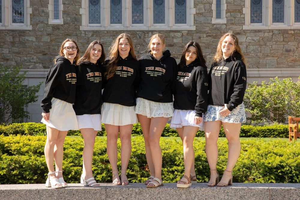 A group wears matching black jackets and white tennis skirts while standing together in front of stone building and garden.