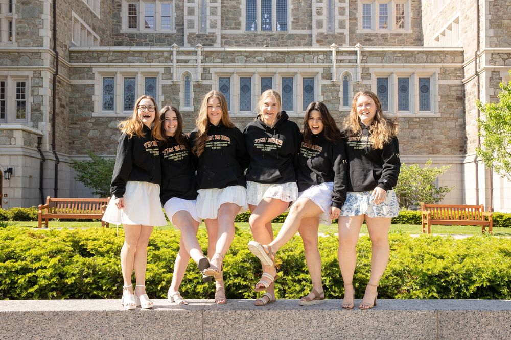 A group in matching black sweatshirts and white skirts pose together in front of a stone building with manicured gardens.