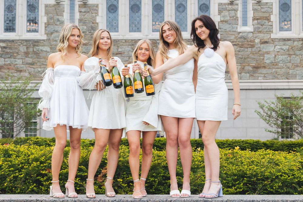 A group in white dresses poses together holding champagne bottles at a celebratory outdoor event.