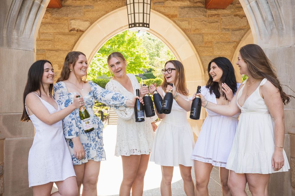 A group in white dresses celebrates with champagne bottles under a stone archway on a sunny day.