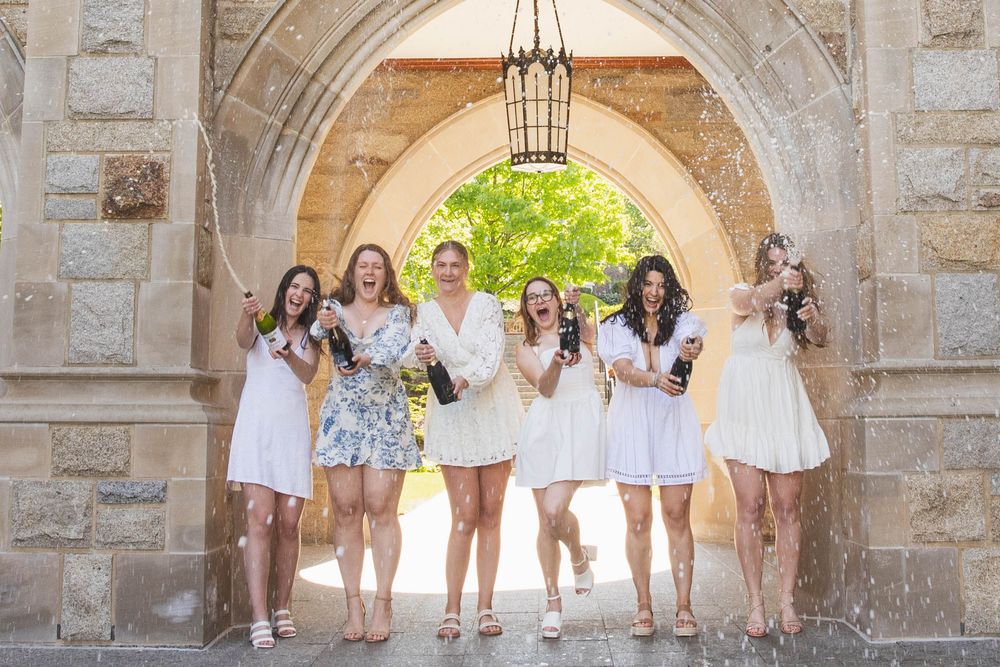 A group in white dresses stands together laughing under a stone archway on a bright sunny day.