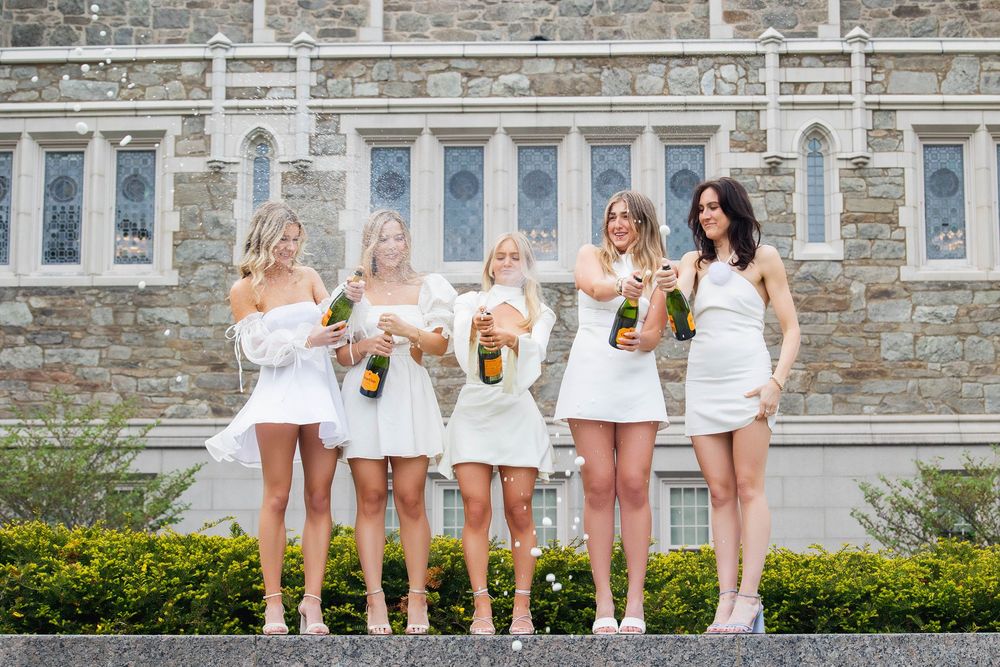 Five people in matching white outfits pose together on steps with drinks in front of a stone building with windows.