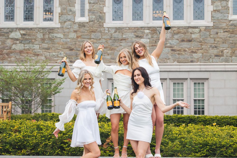 A group wearing white dresses celebrating and posing joyfully in front of a stone building with manicured shrubs.