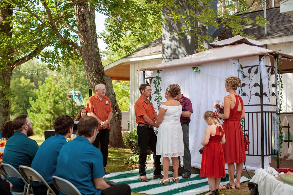 Outdoor garden wedding ceremony with guests gathered around white gazebo surrounded by green trees and florals.