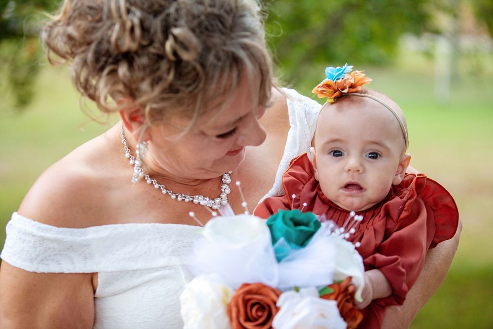 An elegant bride in white off-shoulder dress holding a baby wearing a rust-colored dress with a colorful hair bow.