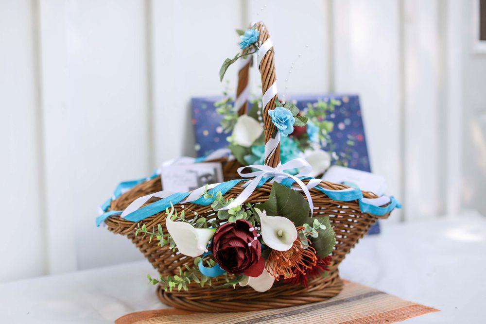 Rustic woven basket filled with artificial blue and burgundy flowers with sage green foliage on white background.