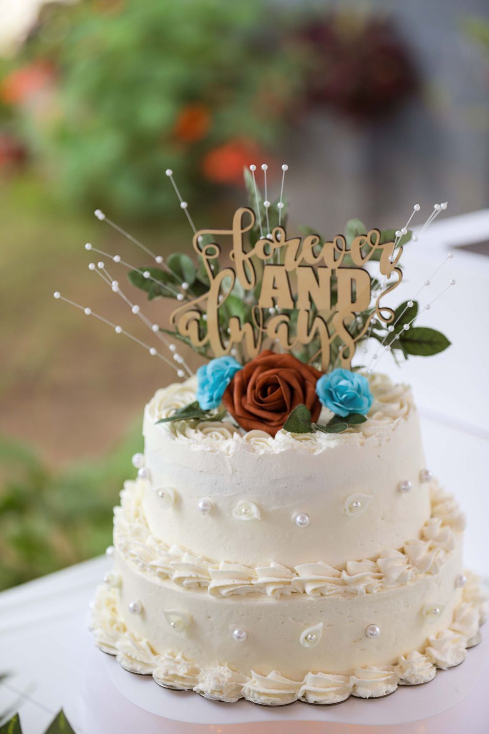 Two-tiered white wedding cake with 'Forever and Always' wooden topper decorated with brown and blue roses and pearls.