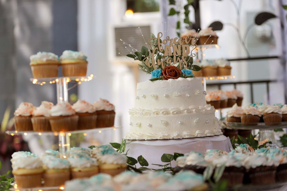 A rustic white wedding cake with greenery decoration surrounded by cupcakes displayed on tiered stands.