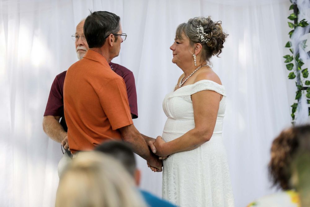 A couple in formal attire stands at an altar during a wedding ceremony with white curtains in the background.