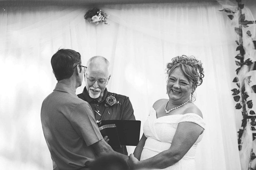 Black and white wedding ceremony photo shows a couple exchanging vows with an officiant at an indoor venue.