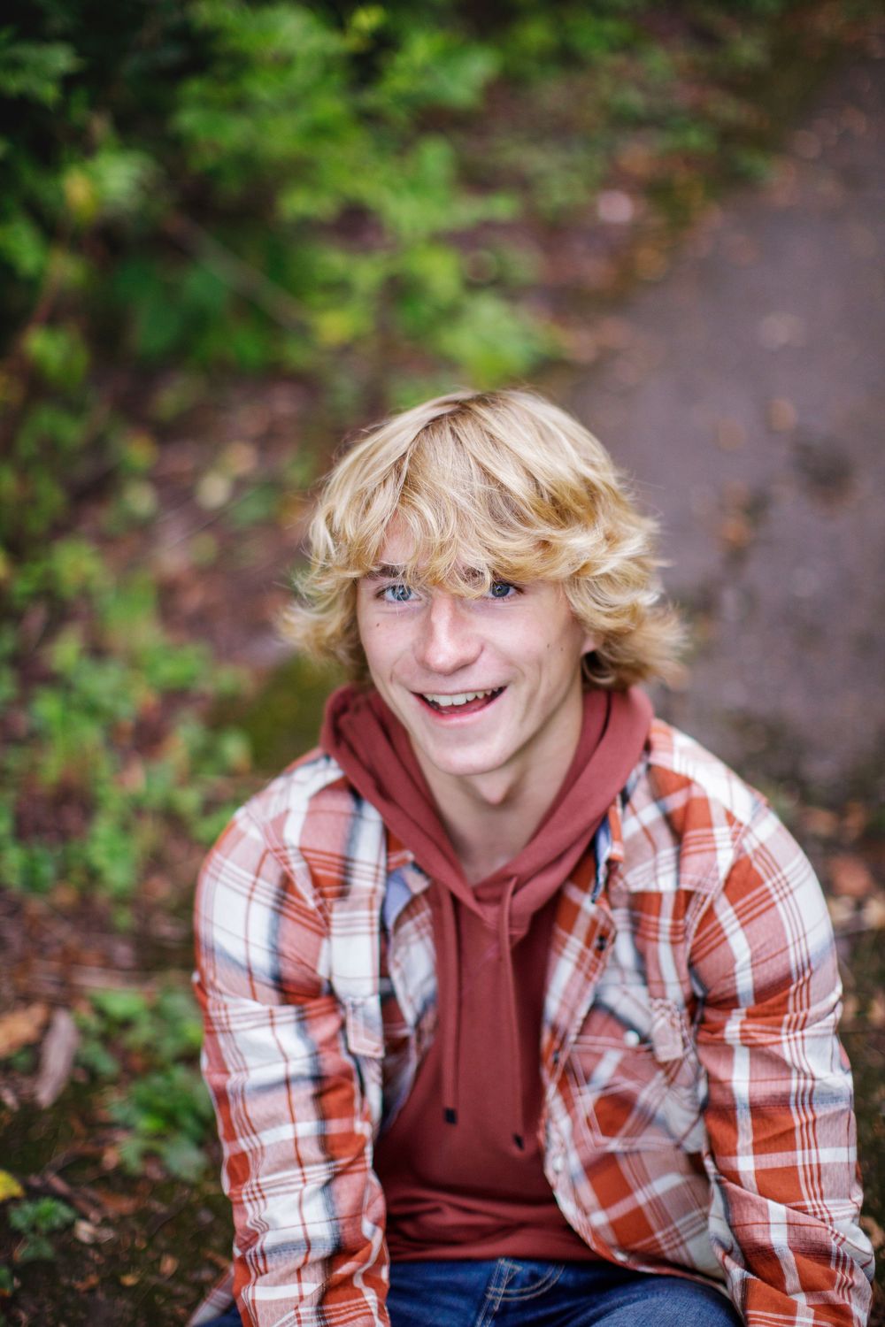 A person in a plaid flannel shirt and burgundy hoodie sits outdoors with autumn foliage in the background.