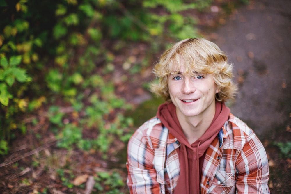 A person with wavy blonde hair wearing a plaid shirt and red hoodie smiles while outdoors among autumn foliage.