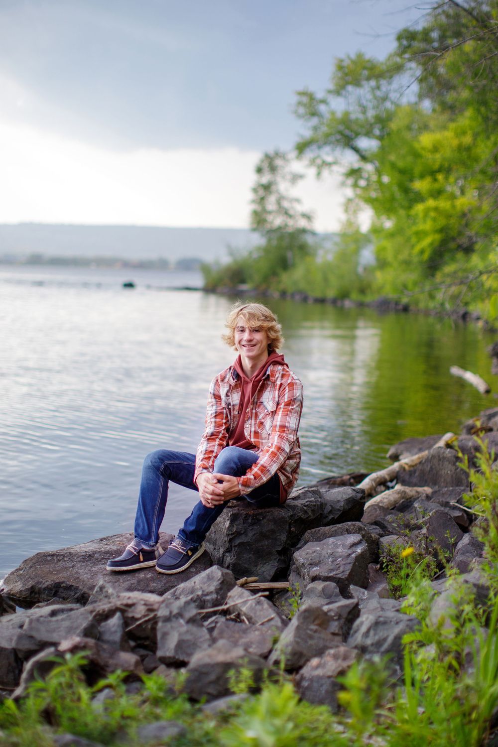 A plaid-shirted figure sits on rocky shoreline next to a serene lake surrounded by green trees on a cloudy day.