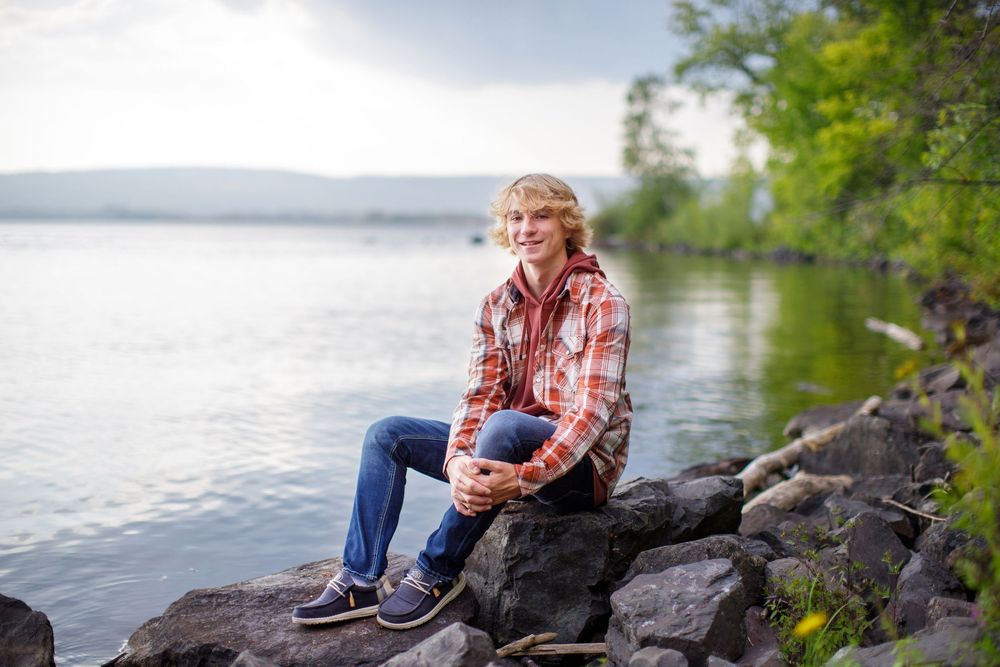 Someone in a plaid shirt sits casually on rocks by a serene lake, surrounded by green trees on a calm day.