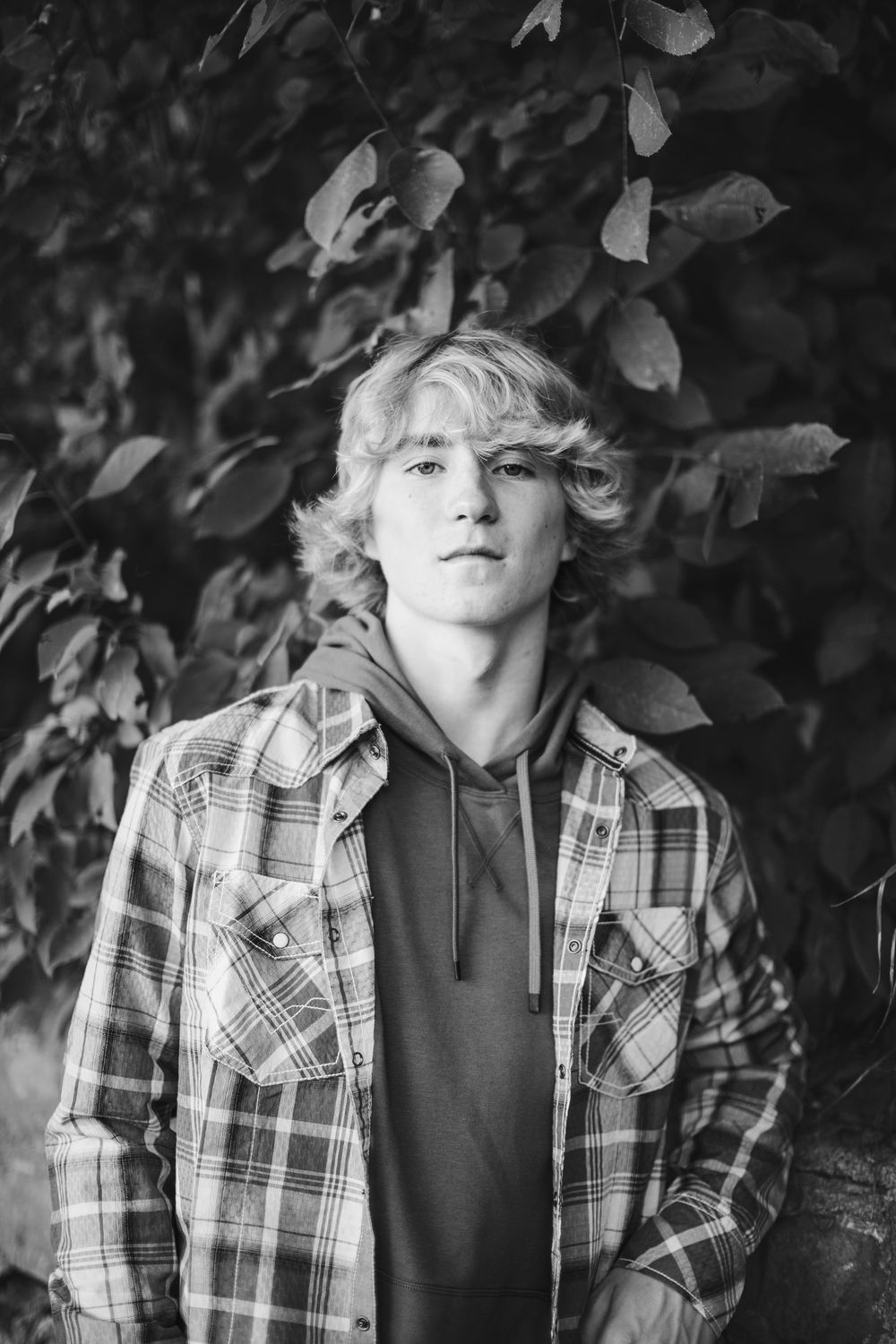 Black and white portrait of someone in a plaid flannel shirt and hoodie against a leafy background.