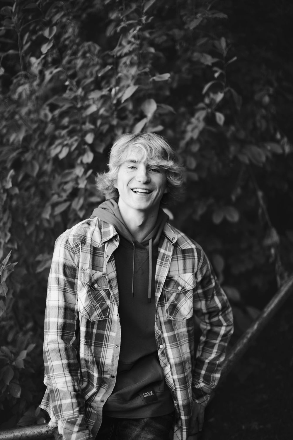 Black and white portrait of a teenager in plaid flannel shirt and hoodie smiling outdoors against foliage background.
