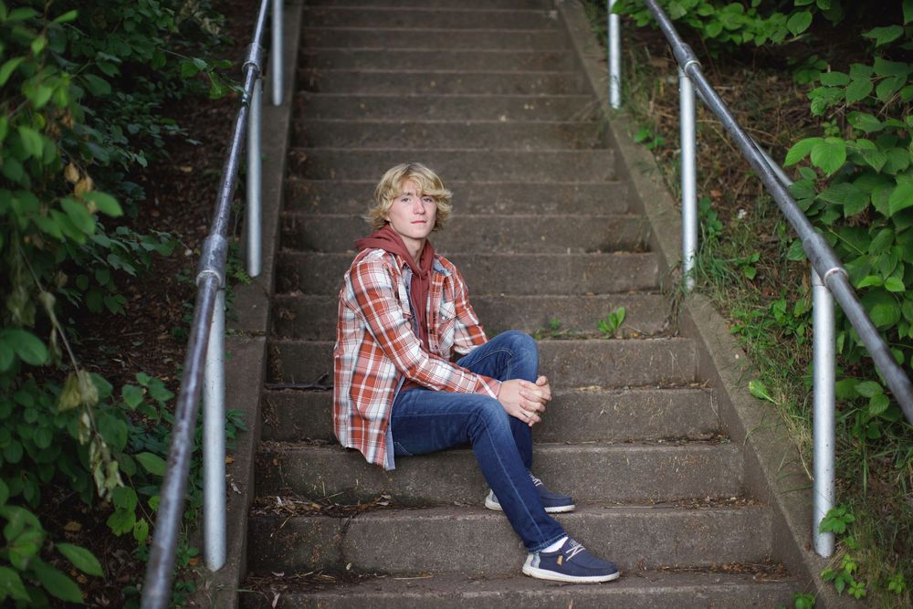 Someone in a plaid shirt and jeans sits casually on outdoor concrete stairs with metal railings surrounded by greenery.
