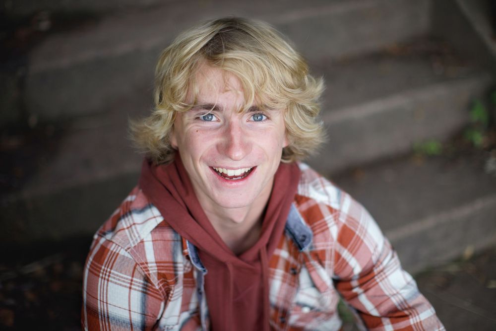 A person with blonde wavy hair wearing a red hoodie and plaid shirt smiles outdoors.