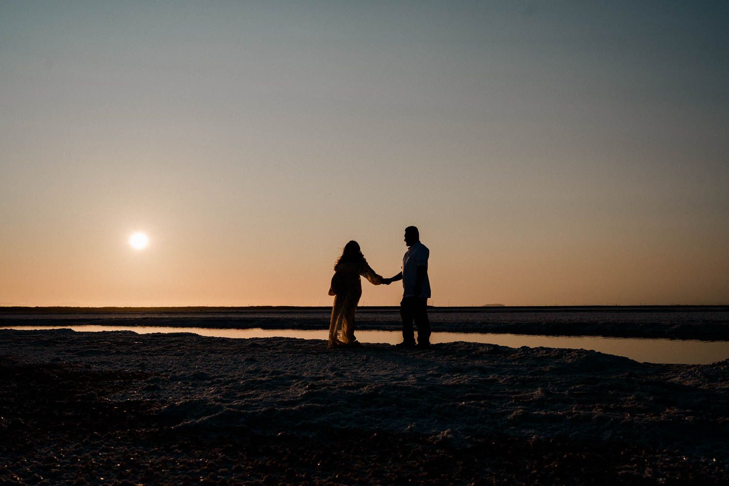 Two silhouettes stand on a rocky shore at sunset while soft waves lap against the coastline in the distance.