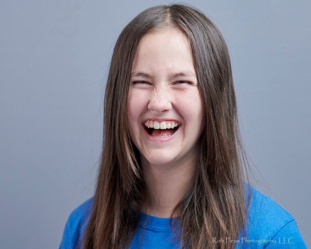 My daughter in the studio laughing with a great big smile on her face. She has long straight hair, a blue shirt, in front gray background.