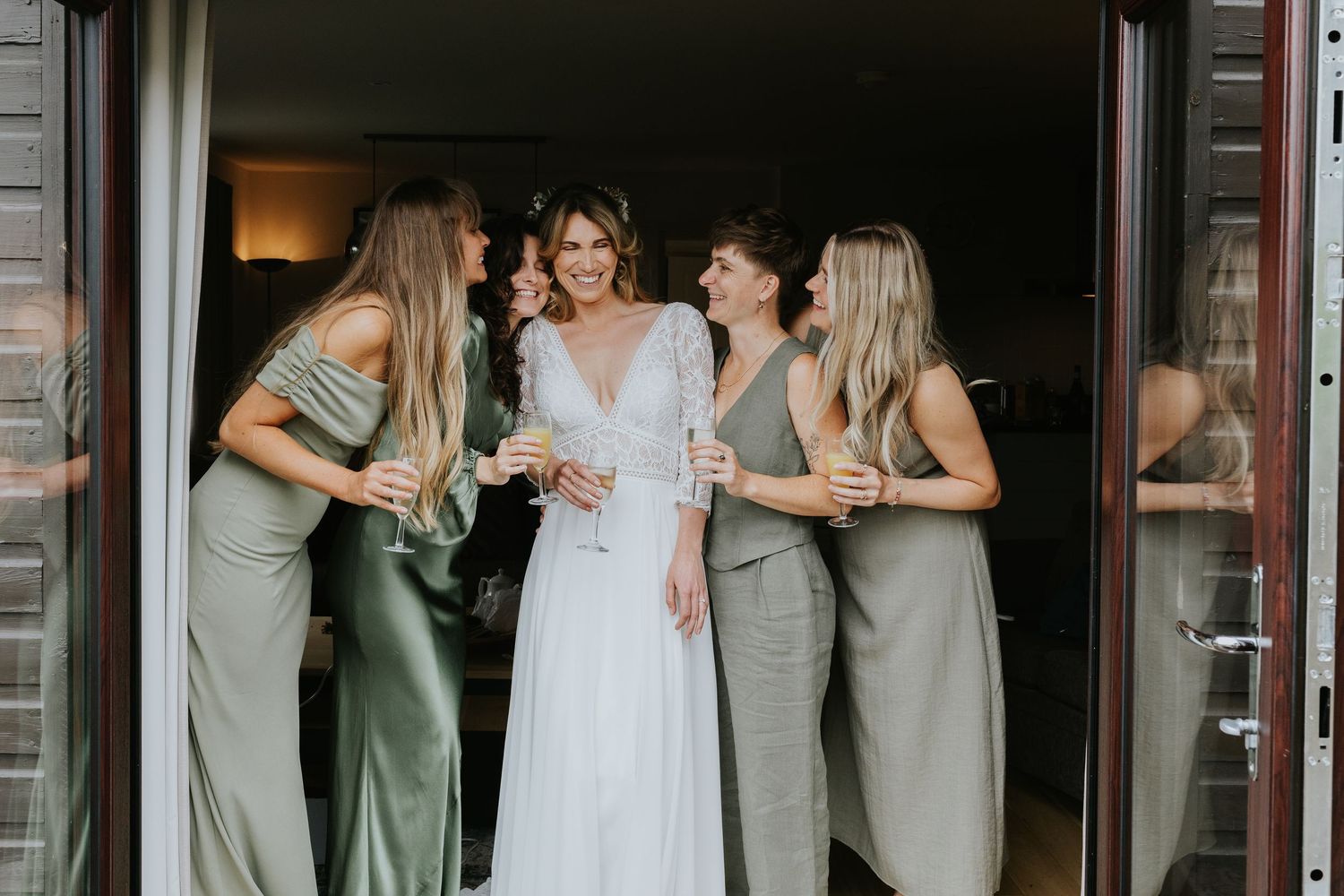 Bride in white gown shares joyful moment with bridesmaids wearing sage green dresses in doorway.