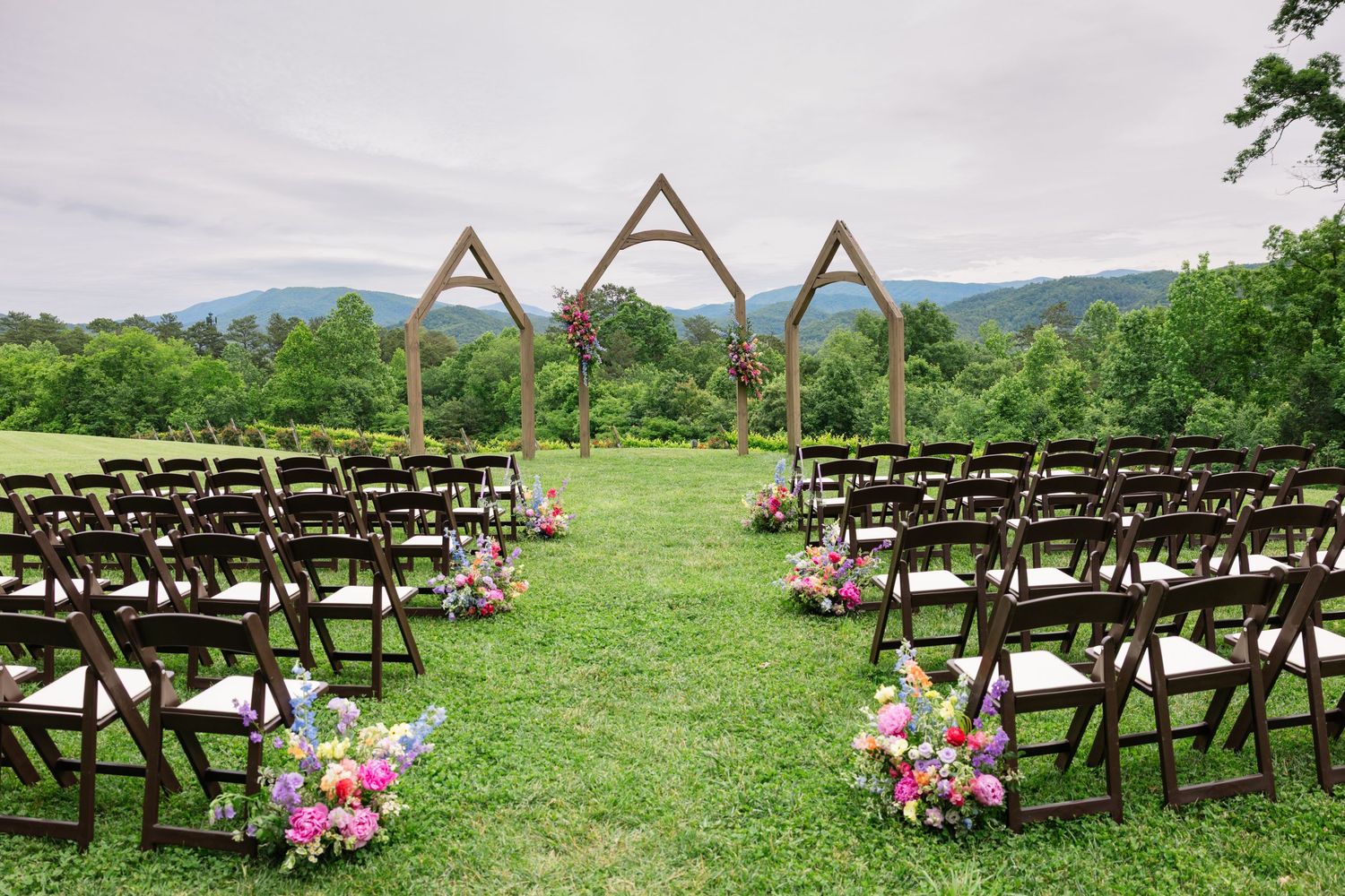 Wooden chairs line a grassy outdoor wedding ceremony site with triangular arch frames and pink floral arrangements.
