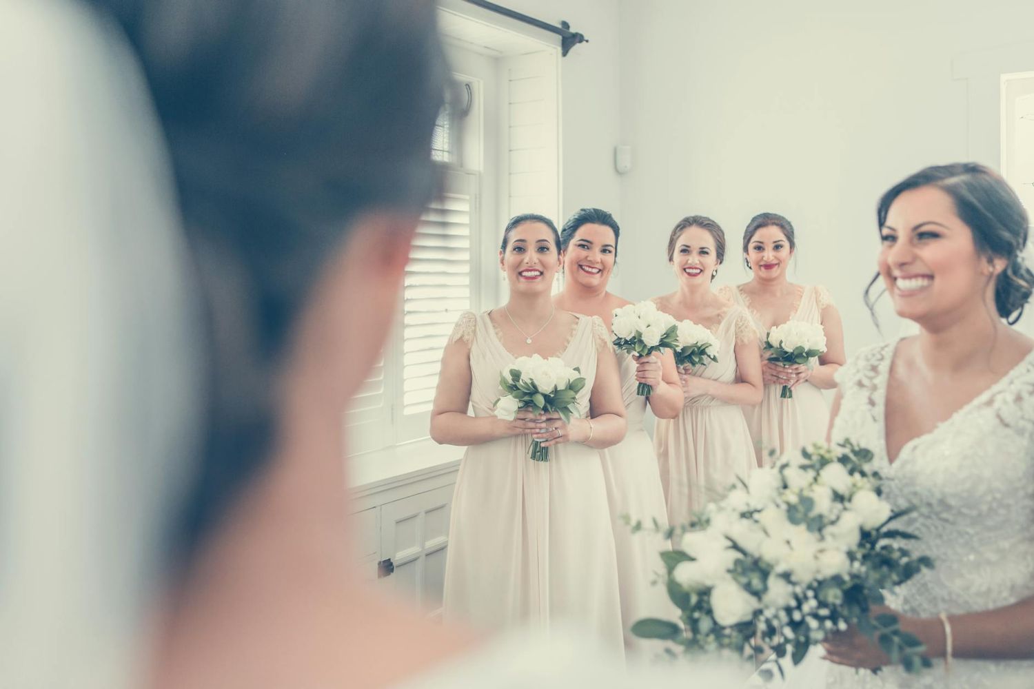 A group in formal attire gathers indoors at a wedding celebration with white floral bouquets and light decor.